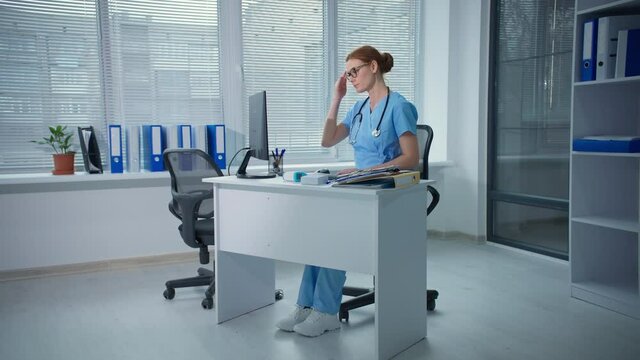 stress at work, overworked female doctor types on a computer keyboard then feels headache while sitting at a table with documents at hospital