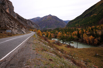 Straight asphalt two-lane road going along the narrow slopes of a high mountain with a beautiful turquoise river flowing below.