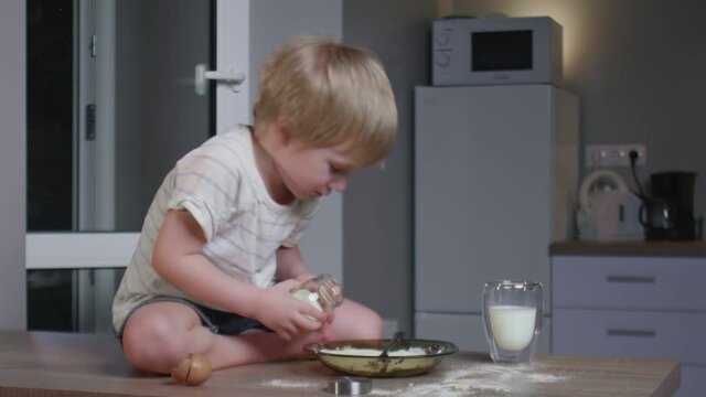 little boy sits on table and pours milk into plate with an egg, mixes and taste it