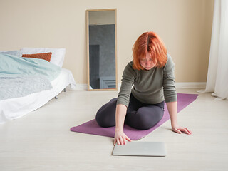 Red hair girl at home in bedroom. Woman sitting on yoga mat and opening laptop. Active lifestyle concept