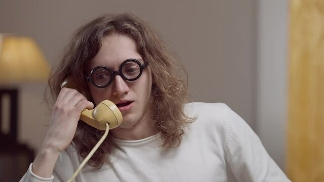 Portrait of genius nerd young man in retro eyeglasses talking on vintage phone indoors. Smart Caucasian student or worker in 1980s or 1990s at home. Lifestyle and intelligence concept.