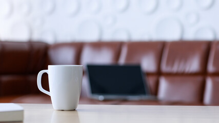 Closeup shot of white coffee mug placed on left side of wooden table in front of brown leather cozy sofa, black laptop computer monitor and white circle shape surface wall in blurred background