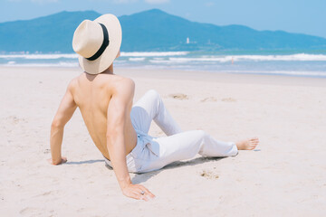 Barebacked young Asian man sitting on the sand and looking at the sea