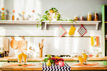 On a wooden tabletop there are a bowl of fruit, glass cups with napkins. In the background is a stylish decorated kitchen interior