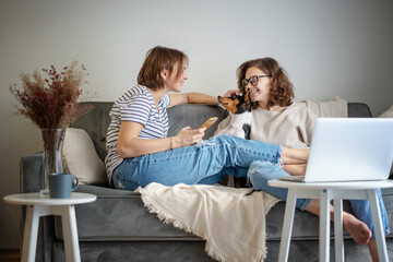 Beautiful happy lesbian couple sitting on sofa at home with pet dog bassengi online chatting using laptop