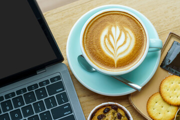 Close-up of Hot coffee latte with latte art milk foam in cup mug and Homemade Banana cup cake with laptop computer on wood desk office desk in coffee shop at the cafe,during business work concept