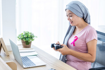 a asian women disease mammary cancer patient playing computer game on laptop with joysticks with pink ribbon wearing headscarf After treatment to chemotherapy in office at home,medicine concept