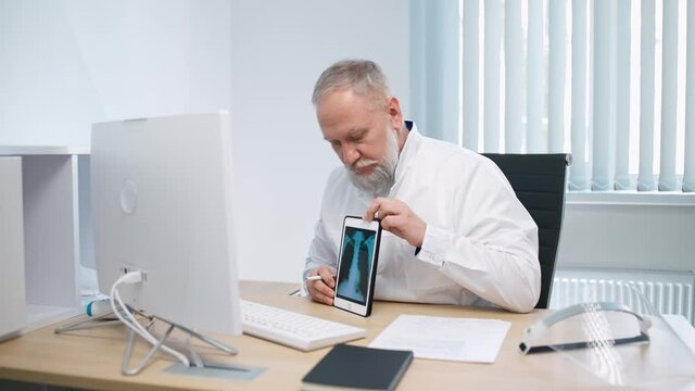Gray-haired male doctor sits at workplace and communicates with patient via video call, the doctor consults the patient online, showing the X-ray image on the tablet screen, modern hospital.