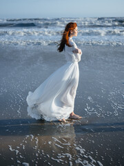 Fototapeta na wymiar A woman in a white dress walks on the wet sand on the shore of the ocean in full growth