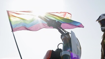 A biker girl sits down on her sports bike against the backdrop of the sun and the LGBT flag waving in the wind.