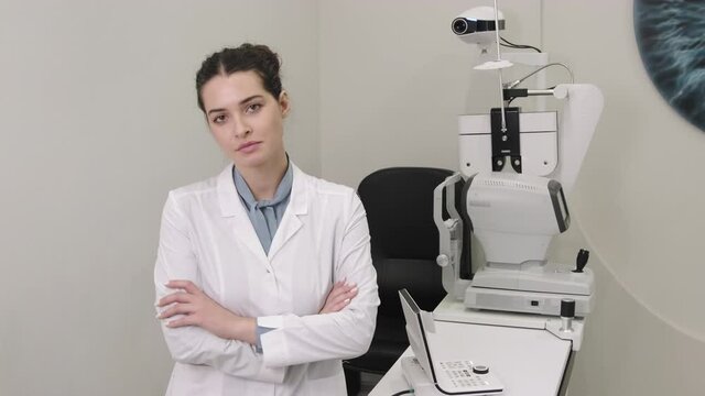 Slow-motion medium portrait of young attractive female optometrist standing with hands folded in office next to modern phoropter used for eye check-up looking at camera smiling