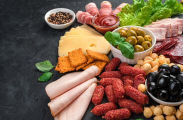 A large assortment of sausage and cheese is sliced on a slate board on a black background.