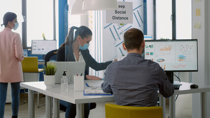 Teamworkers wearing face masks while working at marketing project using computer sitting at table desk in business company office. Coworkers keeping social distancing to avoid infection with covid19