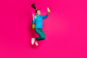 Full length photo of beautiful lady wear blue sweater backpack jumping showing v-sign modern device isolated pink color background