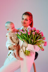 Happy stylish business woman in a light pantsuit with a baby in her arms on a white background with a bouquet of tulips. Mother and child. Shot with colored pink and blue neon light.