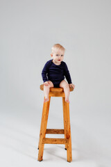 A child sitting on a wooden high chair, isolated against a gray background, a charming baby in a blue bodysuit, looks focused, waiting for his mother to play with him. The concept of childhood.