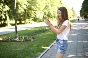 Naklejka premium Girl with a water pistol. Summer fun in the park with a children's water gun. Blonde with long hair in denim shorts and a white T-shirt. Play with water, shoot with water