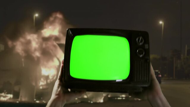 Protestor holding Old Fashioned Television Green Screen in Front of a Burning Barricade Outdoors. You can replace green screen with the footage or picture you want with “Keying” effect in AE. 4K.