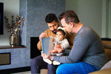 gay couple and their daughter sitting on a sofa reading a story to their little girl. - Powered by Adobe