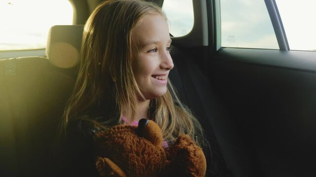 Pretty girl with teddy bear looking out window during summer car trip, lifestyle. Sun light on the face of passenger. Facial emotions of little traveler. Happy traveling family concept.