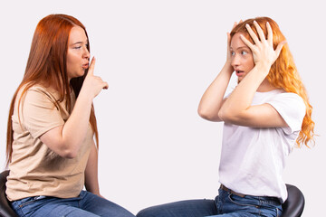 Young pretty red haired girl making silence gesture, hush with finger on lips to her frightened, scared friend, asking to be quiet. Silence or secret concept on gray background, free space for text