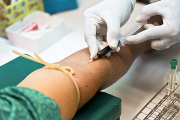 Doctor or nurse hands in medical white gloves using needle syringe drawing blood sample from patient arm in hospital.