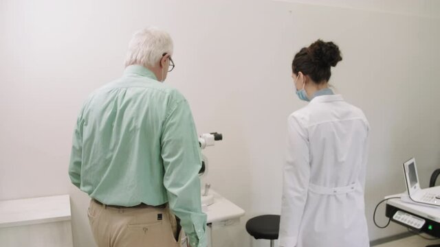 Medium shot of caucasian senior man entering modern doctor office greeting female ophthalmologist, sitting next to eye checkup machine getting ready for eyesight testing