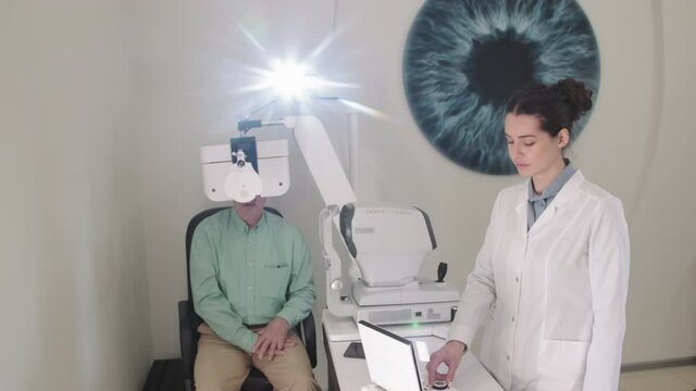 Medium shot of young female ophthalmologist using ophthalmic eye chart projector while senior man doing eye checkup at modern office