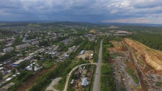 Aerial view of small Provincial industrial city. On the outskirts of the city there are low houses, garages and quarries. Cars drive along the road. Summer evening, in the sky doe clouds and blue clou