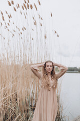 a beautiful girl with brown hair in a beige dress stands in the tall reeds by the water
