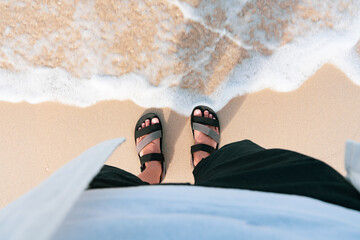man in Black slippers  flip flop feet standing on the beach with a wave of foaming gentle beneath...