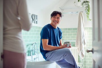 Mature Asian Couple Wearing Pyjamas Sitting In Bathroom Enjoying Morning Hot Drinks Together