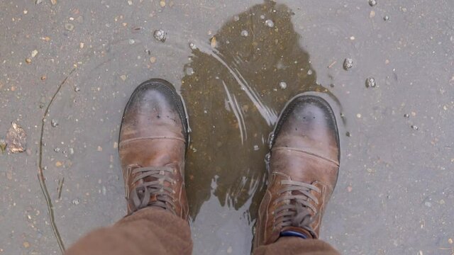 Staying in a puddle on an asphalt surface. POV or FPV of men boots wading in the water on wet autumn day at the urban street. First person view of feets splashing water slow motion shot.