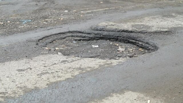 Pit in the wet asphalt caused by bad weather, traffic and poor maintenance. Slow motion panorama of a road hole.