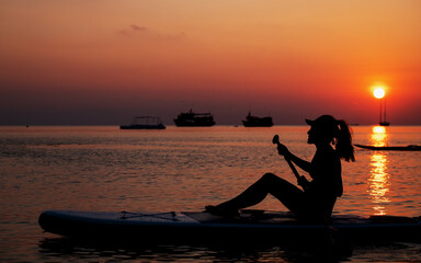 Silhouette image of a young woman sitting on stand up paddle board in the sea before sunset