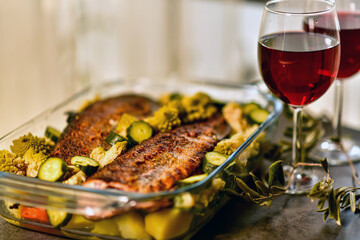 two glasses of red wine and a large glass dish with baked fish and vegetables
