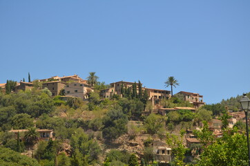 View of the village, which is located on the top of the mountain. The village consists of brown houses that glow in the rays of the summer sun. There are many green plants around.