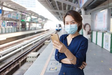 Asian lady who has short hair play smartphone while waiting train at station for travel and wears medical face mask as new normal lifestyle.