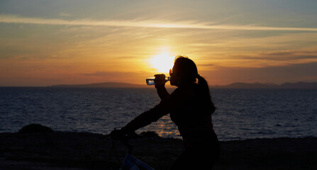 Backlit, woman drinks water leaning on her bicycle, at sunset