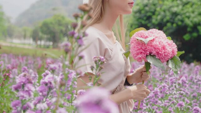 a light skin long blnde hair woman slowly walks in beautiful purple flowers field in summertime, young attractive girl holding a bright pink bouquet  while strolling in flower farm on weekend, 