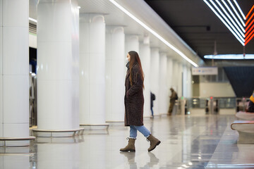 A full-length photo of a lady in a medical face mask to avoid the spread of coronavirus who is going on the subway platform. A girl in a surgical mask is keeping social distance on a metro station.