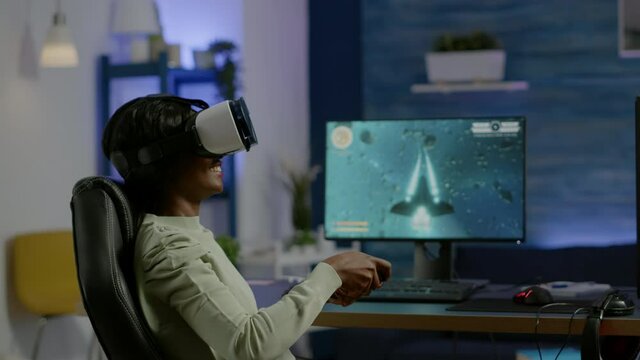 Black woman gamer playing video game at powerful computer late night wearing vr headset. Excited player using wireless controller for virtual tournament gaming space shooter at home