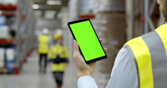 Back view of male worker in warehouse wearing safety vest standing near shelf using cellphone