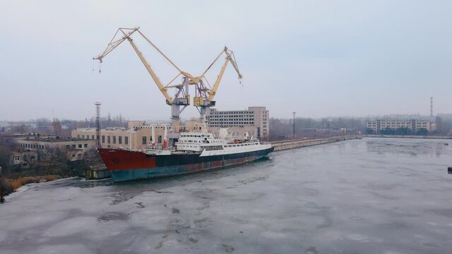 Aerial view of shipbuilding yard with huge cargo crane and old rusty vessel standing on water with melting ice and floes. Concept of transport logistics and maritime in winter