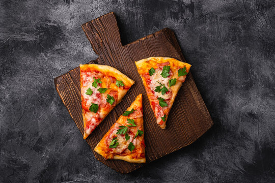 Hot pizza slices with mozzarella cheese, ham, tomato and parsley on brown wooden cutting board, stone concrete background, top view