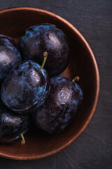Fresh ripe plum fruits with water drops in wooden bowl on dark stone background, top view macro