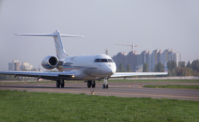 
A private twin-engine jet aircraft moves along an airport taxiway with its headlights on against a clear sky