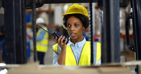 Young african woman standing near forklift and using walkie-talkie in warehouse