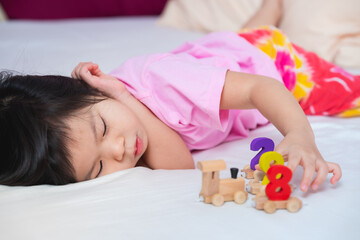 Obraz na płótnie Canvas Cute Asian girl is lying on a soft white mattress. Child play wooden toys, train number figures happily. Children feel safe when stay with family. Kids wearing a pink dress is 4 years old. Relax time.