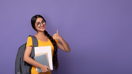 Smiling indian student wearing eyeglasses holding notebooks showing thumbs up